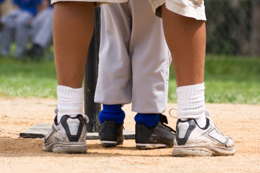 Legs of a coach and child with a t-ball base. Coach is teaching the player how to bat and play the game of baseball.