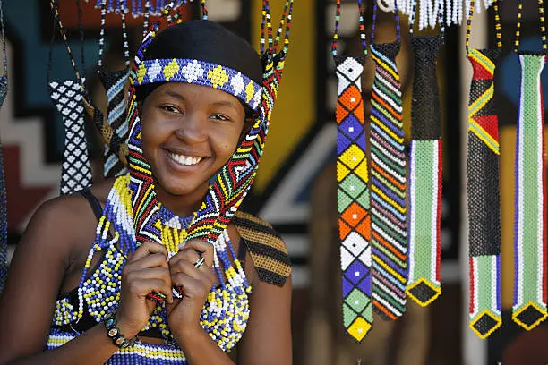 "A pretty young Zulu girl poses amongst beaded souvenirs that she has hand made and selling to visitors to this area of Kwazulu-Natal, in South Africa."