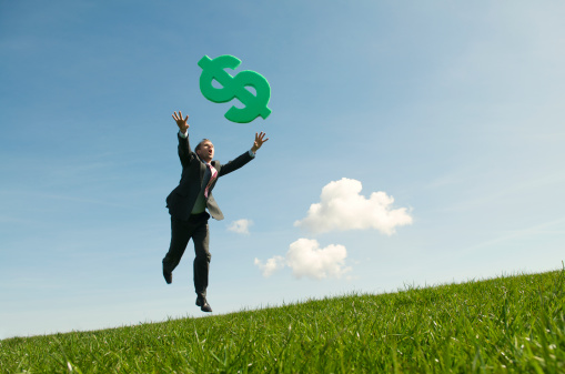 Businessman leaps above a green meadow chasing a big green foam dollar sign