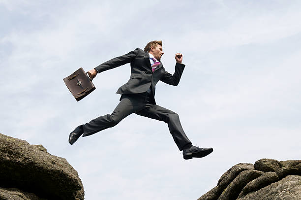 Businessman Jumping Outdoors Between Rock and Hard Place White Sky Young man businessman jumping outdoors with his briefcase between two rocks against white background sky leap of faith stock pictures, royalty-free photos & images