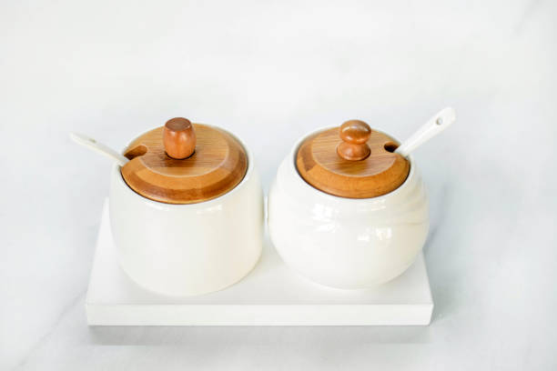 A salt shaker and a sugar bowl in white with wooden lids stand on a white table. Elements of decor in a modern interior. Modern White Tableware.