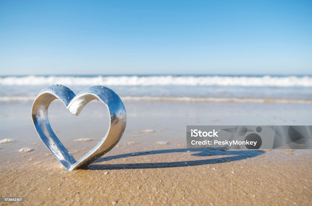 Romantic Honeymoon Silver Heart on Beach in Sand with Waves Silver heart stands outdoors on the beach in textured brown sand washed by gentle waves Beach Stock Photo