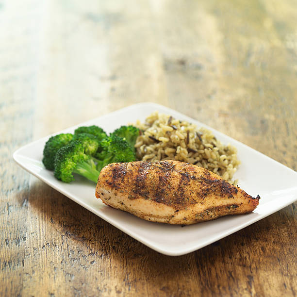 Herbed, grilled, skinless chicken breast on rectangular plate with broccoli, and long grain and wild rice.  Shot with shallow focus on chicken breast shot on rustic wood table.  Plenty of room for copy.