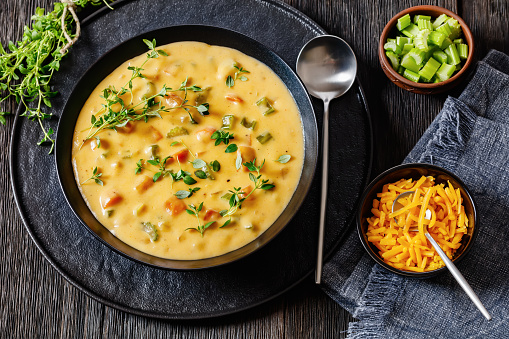 Beer cheese soup with vegetables and thyme in black bowl on dark wooden table with spoon and ingredients, horizontal view