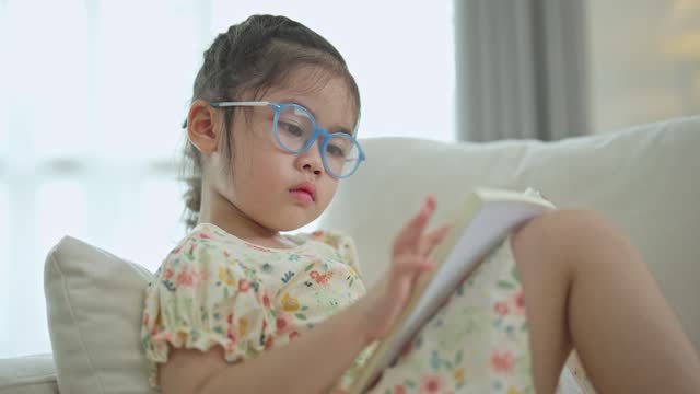 Joyful happy asian child baby girl smiling and reading book while sitting on couch sofa in living room at home. Girl relex reading book smile at sofa In the house. Back to school concept.