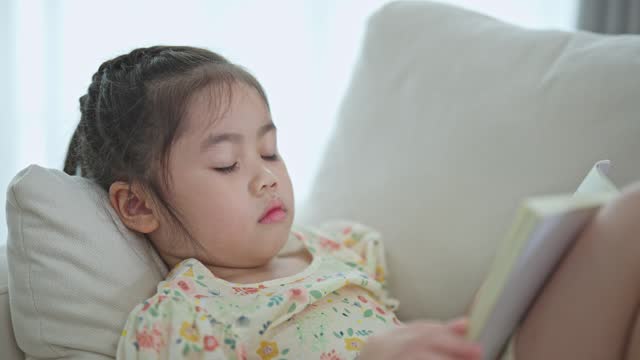 Joyful happy asian child baby girl smiling and reading book while sitting on couch sofa in living room at home. Girl relex reading book smile at sofa In the house. Back to school concept.