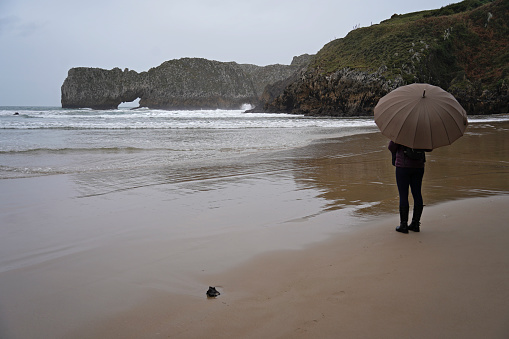 One person enyoing the beauty of a wild beach in cold and rainy weather, protected by an umbrella