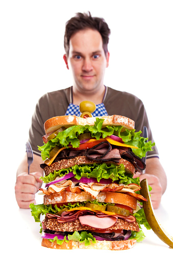 Photograph of a huge deli sandwich, Dagwood Style, with man in background wearing a checkered napkin and big eyes; man is blurred and sandwich is in focus; copy space 