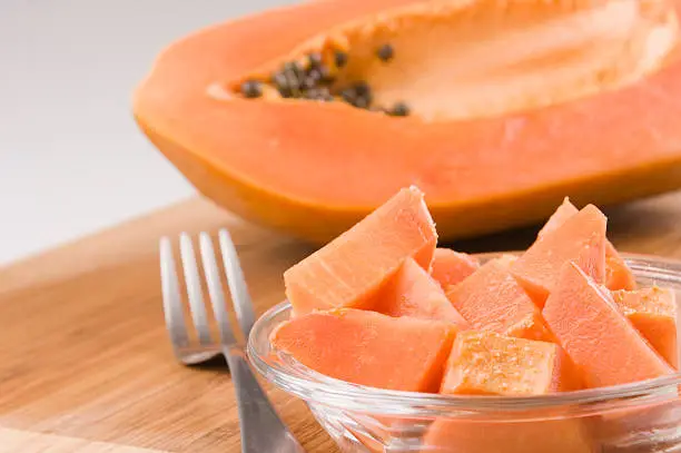 Serving of small chunks of papaya fruit on cristal bowl, fork on the left and everything placed on a bamboo cutting board with half papaya in the back.