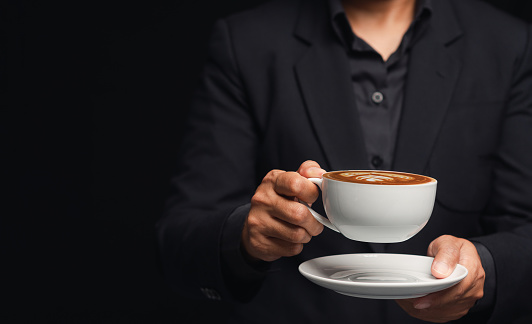 Businessman in a suit is holding a cup of coffee while standing on a black background. Close-up photo