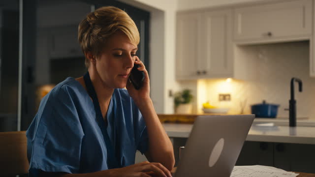 Tired Woman In Medical Scrubs Talking On Mobile Phone Working Or Studying On Laptop At Home At Night