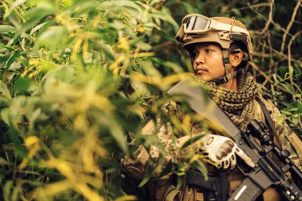 Photo of American soldiers resemble rangers from the Army. Carrying an M16 rifle and a pistol, hiding in the bushes wearing camouflage uniforms and vision goggles, ready to fight enemies on the battlefield.