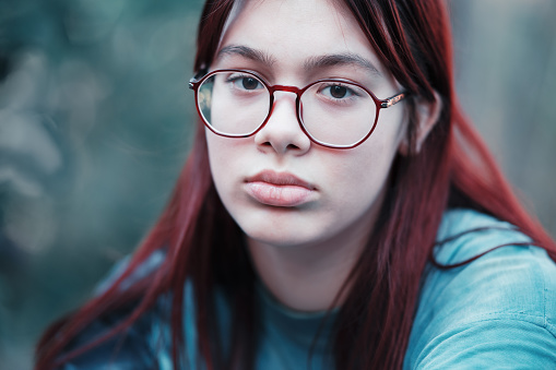 Portrait of a sad girl with glasses. A thoughtful red-haired teenage girl in a blue T-shirt looks at the camera..