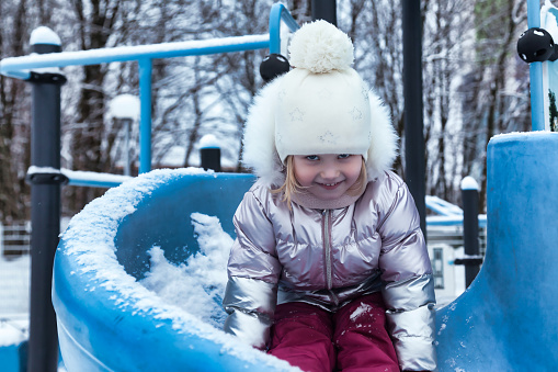 Happy little kid girl on slide in winter snowy playground in warm wear, smiling looking at camera. Adorable joyful child playing outdoors. Concept of wintertime and childhood. Copy ad text space
