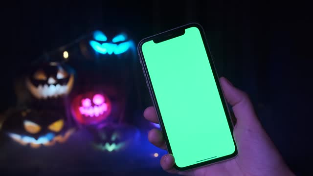 Phone with green screen held by hand on a background of many flashing multi-colored neon lights carved pumpkins
