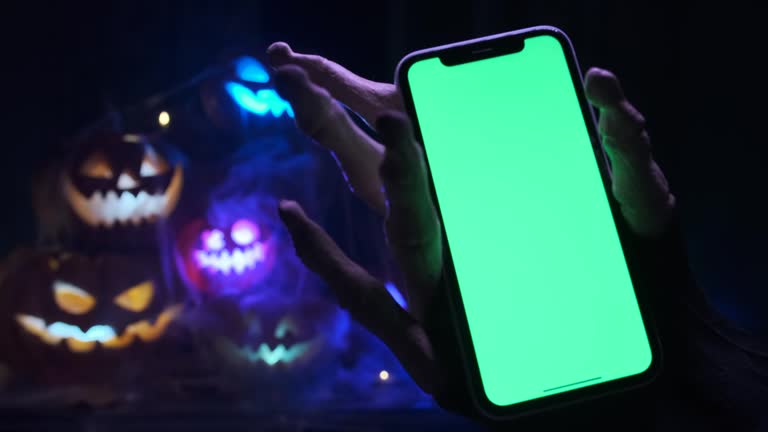 Monster hand holding phone with chroma key against background of pile flashing Jack-o'-lanterns glowing carved pumpkins