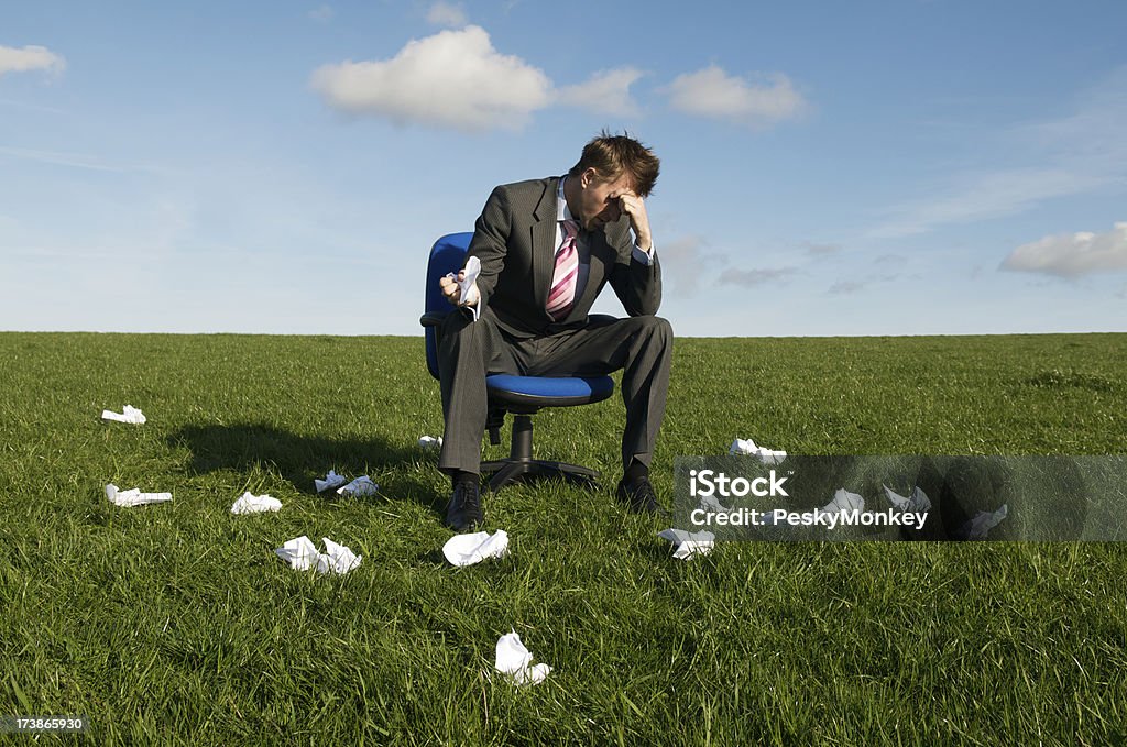 Frustrated Businessman Sitting in Meadow with Rejected Crumpled Paper "Frustrated businessman sits in a green meadow on his desk chair, the grass around him littered with crumpled paper" Businessman Stock Photo