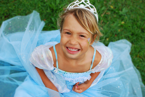 Little girl dressed as a pretty blue princess with tiara sitting on the grass.
