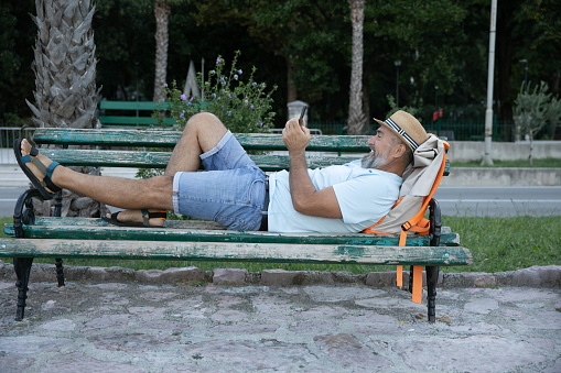 A gray-haired senior man lying on a bench by the sea is smiling and looking at his smartphone. Attractive smiling people enjoying nature and the beauty of nature. Older people and modern technology