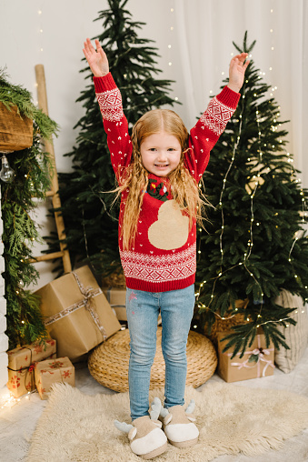 Child girl in stylish Christmas sweater with deer and comfortable home slippers near Xmas Tree. Happy kid stands near Christmassy gift, presents for New Year. Decorated interior with garlands, lights.