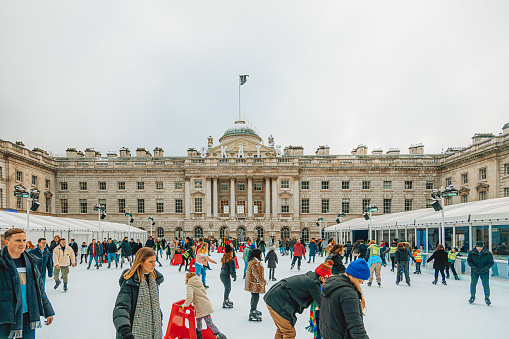 A heartwarming scene captures the joy of people skating on the seasonal ice rink in the picturesque courtyard of Somerset House, located in London's historic Strand district at Christmas Time. The neoclassical elegance of Somerset House, framed by Georgian architecture, serves as a delightful backdrop to the holiday festivities. This remarkable 18th-century structure now thrives as a hub for cultural events, including The Courtauld Institute, and in the summer, the courtyard transforms into The Edmond J. Safra Fountain Court, complete with beautiful fountains.