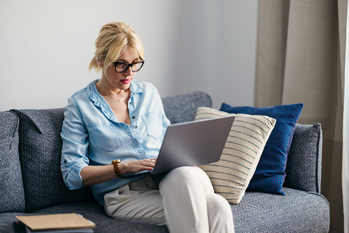 A pensive Caucasian entrepreneur with glasses using her laptop while sitting on the sofa in the living room.