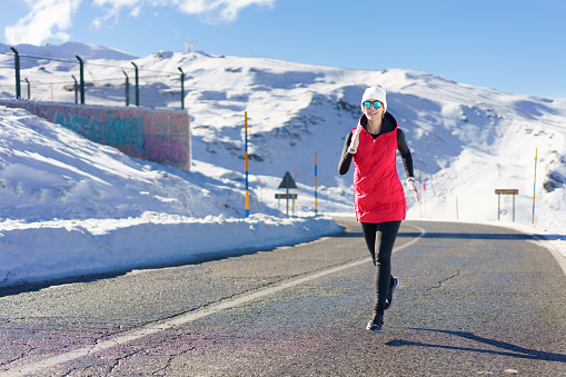 Full body of young female in red vest beanie cap, polarized sunglasses looking at camera while jogging on asphalt road near snow against snowy mountain and blue sky in daylight
