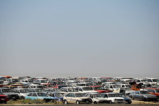 Long shot looking towards a large collection of wrecked cars in a junkyard. Cars are in the bottom third of frame against a heat hazed clear sky.