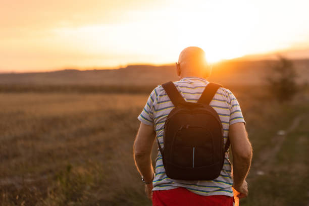 A senior man, carrying a sleek black backpack, leisurely strolls through the peaceful serenity of nature, enjoying a tranquil walk amidst the great outdoors.
