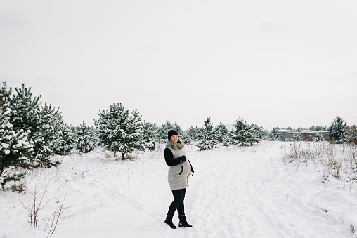 Young pregnant woman wearing warm clothes having fun on a beautiful winter snowy day. Spending pregnancy time in a winter mountain resort. Female walk in park or forest. Winter holidays.