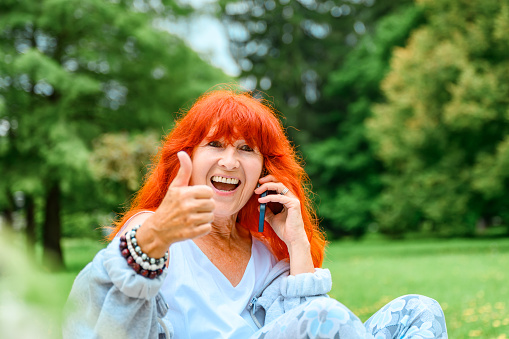 Senior woman with long orange hair talking on a smart phone in a public park and showing her thumb up while smiling and looking at camera.