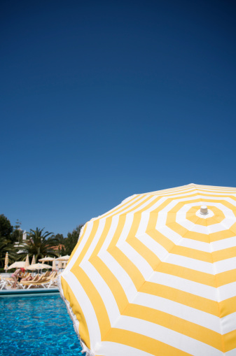 Bright yellow and white striped sun umbrella dominates the foreground of a warm poolside scene