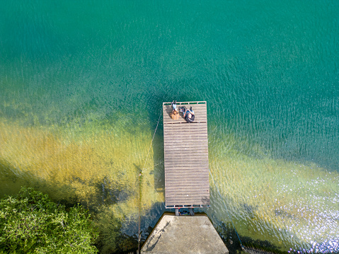 An awe-inspiring aerial view of a wooden pier extending gracefully towards the inviting, clear water of a serene lake. The tranquility of the scene and the natural beauty of the pristine sand and water create a captivating and peaceful visual. Aerial Serenity: Wooden Pier Leading to Clear Water Lake. High quality photo