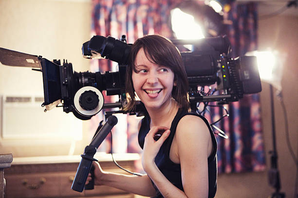 Laughing Woman with Camera "A young woman operating camera on a video shoot. Camera has a 35mm adaptor on it. Shot on a real set at high iso, some noise, view at 100%.More filmmaking images:" film director stock pictures, royalty-free photos & images