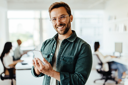 Young designer using a mobile phone in an office. Happy business man looking at the camera while standing in an open plan office.