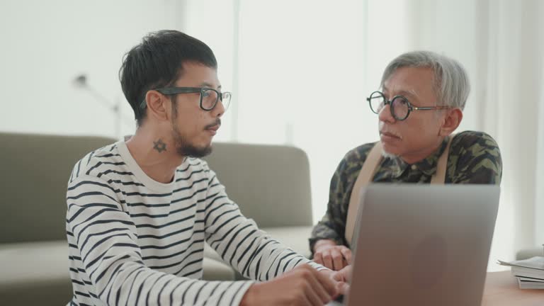 Family Support in the Digital Age: Asian Man Freelancer Talks and Works Alongside Senior Father in Cozy Home Environment.