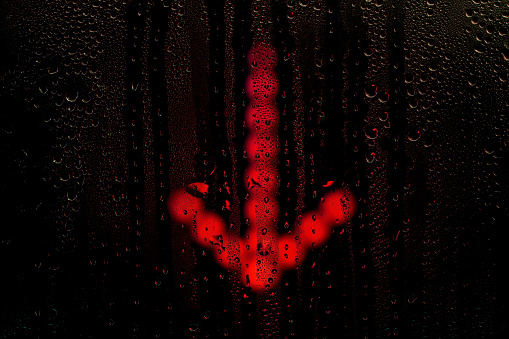 Glowing arrow sign pointing down behind wet glass. Raindrops on the window. Background on the theme of depression and recession.
