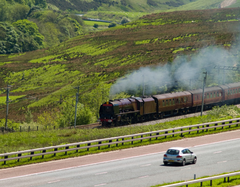 A Princess Coronation (Duchess) class express steam locomotive steaming fast up the west coast main line on the fringes of the English Lake District. A car travelling in the opposite direction is on the adjacent M6 motorway.