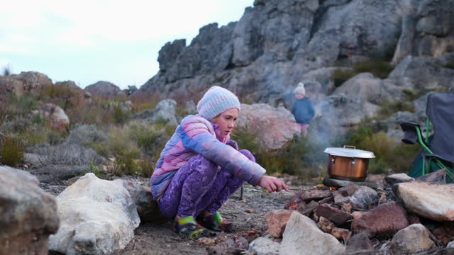 Young girl keeps the camp fire alive with small branches and leaves