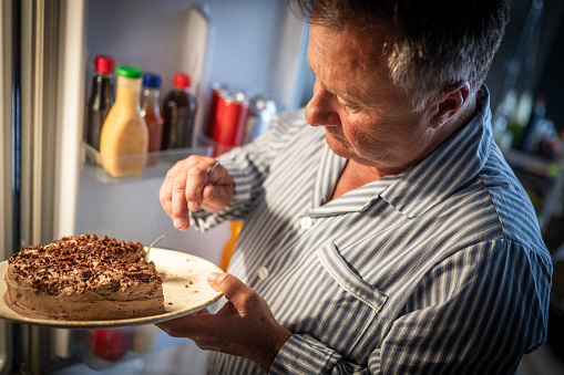 Side view of a mature white man in pajamas eating cake in front of an open refrigerator at night.