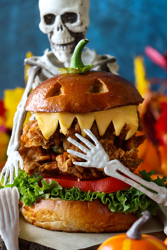 Stock photo showing close-up view of Halloween party food ideas. Homemade, snarling monster designed, fried chicken burger served in brioche bread bun with battered chicken breast on a bed of lettuce and a tomato slice, and topped with processed cheese slice teeth, on a wooden platter surrounded by artificial autumnal fallen leaves against a mottled blue background.