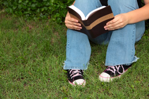 Teenager holding and reading a Bible outdoors in the grass. Horizontal image would be good for Christian or religious use.