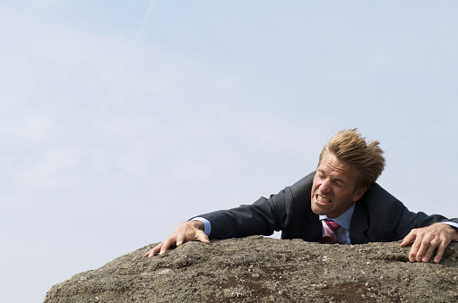 Businessman struggling to the top of a rock, wind blowing his hair, only the sky above him