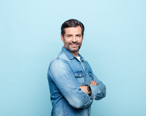 Handsome mature man wearing denim shirt standing with arms crossed against blue background and smiling at camera. Studio shot.