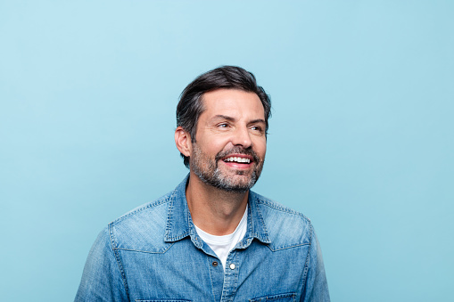 Handsome mature man wearing denim shirt standing against blue background, looking away and smiling. Studio shot.