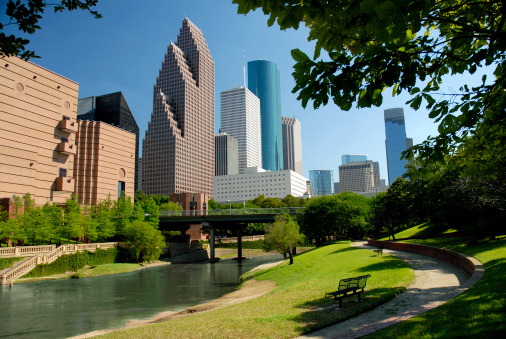 Modern skyscrapers of Houston, Texas are seen in this horizontal color image across a waterway flowing through a city park.  A perfect, clear blue sky is a backdrop to this afternoon cityscape.  Tops of the buildings are somewhat outlined by tree branches.  A park bench and walking trail are included in the foreground.