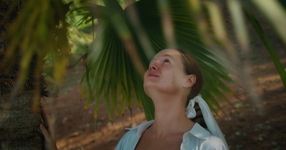 Traveler woman enjoys a walk in tropical garden jungle forest with palm trees. Close-up of girl face between green palm branches. Slow-motion. Immersive travel.