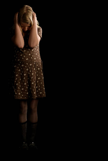 Terrified little girl covering his head while standing in the dark and looking down.