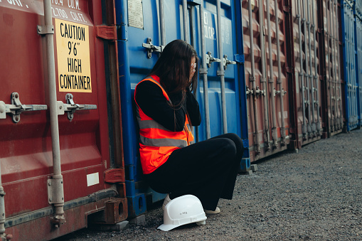 Frustrated and tired engineer woman sitting on ground in shipping container terminal with white helmet. Crisis, construction problem and lost job.