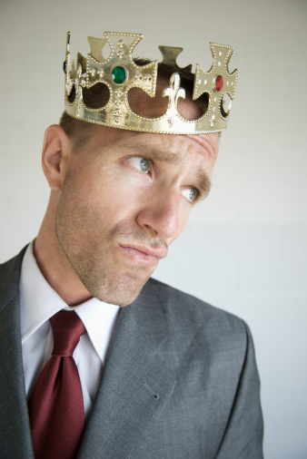 Handsome businessman is holding party crown on stick, looking at camera and smiling, on gray background